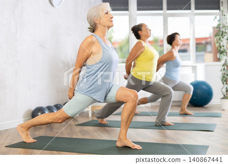 Old woman practicing warrior pose of yoga in training room during fitness classes Old woman practicing warrior pose of yoga in training room during fitness classes 140867441