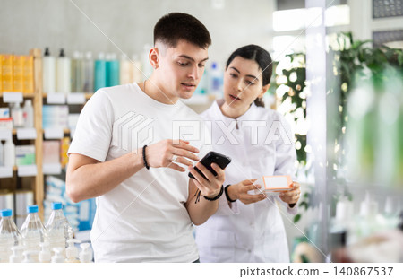 Guy customer looking for something in a drugstore, checking the list on her smartphone. Female pharmacist offers him medicines Guy customer looking for something in a drugstore, checking the list on her smartphone. Female pharmacist offers him medicines 140867537