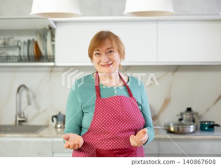Elderly smiling lady in apron stands in clean kitchen and comes up with menu for lunch 140867608