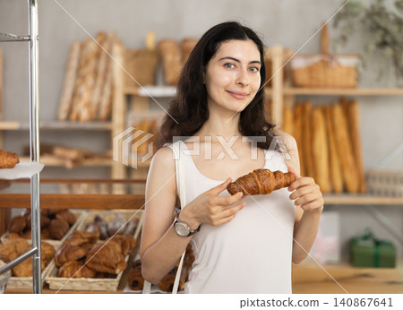 Young female customer stands with croissant in hands near window of bakery. Young female customer stands with croissant in hands near window of bakery. 140867641