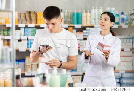 Handsome young man choosing box with pills in pharmacy, scan QR code on label and professional pharmacist woman helping and offering supplements at drugstore 140867664