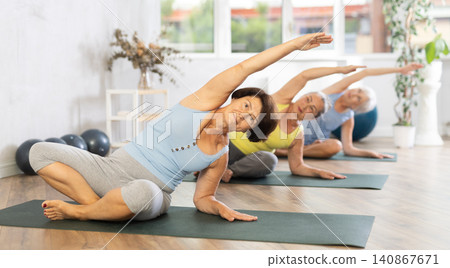 Elderly woman in company of hatha yoga lovers performs half-lotus pose, Ardha Padmasana Elderly woman in company of hatha yoga lovers performs half-lotus pose, Ardha Padmasana 140867671