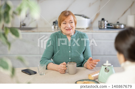 In kitchen at table, elderly smiling woman communicates with friend and drinks coffee or tea In kitchen at table, elderly smiling woman communicates with friend and drinks coffee or tea 140867679