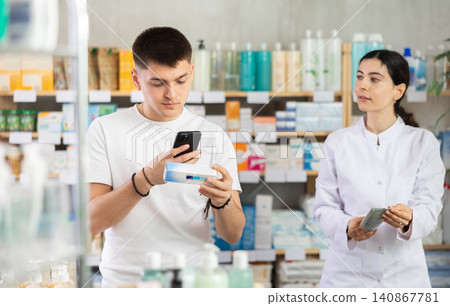 Guy in pharmacy scans QR code on antifungal ointment, pharmacist in background Guy in pharmacy scans QR code on antifungal ointment, pharmacist in background 140867781