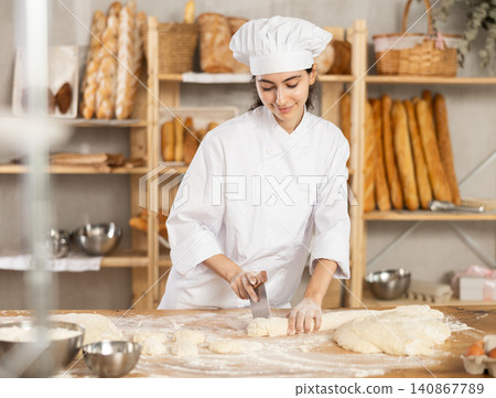 Young female baker cutting dough with scraper Young female baker cutting dough with scraper 140867789