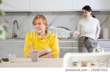 Elderly sad woman sits at table in kitchen and listens to insults.Concept stream of abuse Elderly sad woman sits at table in kitchen and listens to insults.Concept stream of abuse 140867810