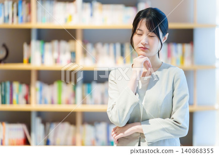 A woman with a troubled expression stands in front of a bookshelf; she is a writer, novelist, and essayist. A woman with a troubled expression stands in front of a bookshelf; she is a writer, novelist, and essayist. 140868355