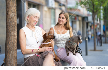 Elderly woman and her granddaughter are sitting on a bench and holding their favorite dachshund dogs Elderly woman and her granddaughter are sitting on a bench and holding their favorite dachshund dogs 140868521