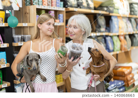 Girl and elderly woman choosing dog food together with dachshund in pet store Girl and elderly woman choosing dog food together with dachshund in pet store 140868663