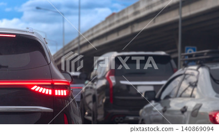 Low angle shot showing the rear of a black car stuck in dense traffic During the day. Turn on brake light. Vehicles are lined up on the highway road beneath a massive concrete overpass structure. 140869094