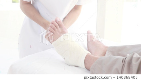 Holding patient's white bandaged right foot, clinician in white uniform using metal tool in clinic Holding patient's white bandaged right foot, clinician in white uniform using metal tool in clinic 140869403