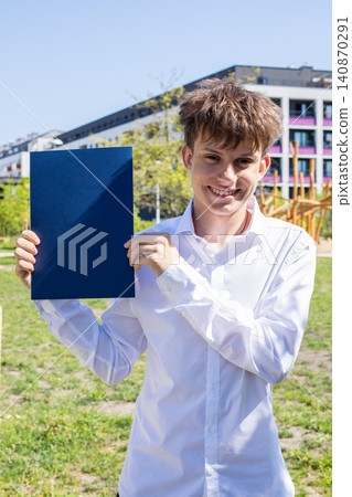 Smiling caucasian teenager holding blank navy blue degree certificate cover outdoors. Concepts of school graduation, success and education. Smiling caucasian teenager holding blank navy blue degree certificate cover outdoors. Concepts of school graduation, success and education. 140870291