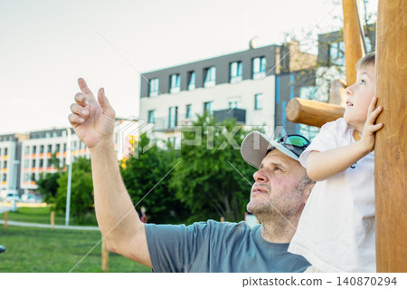A mature father or grandfather points to the sky, showing something to his small boy on a wooden city playground. Family generations together. 140870294