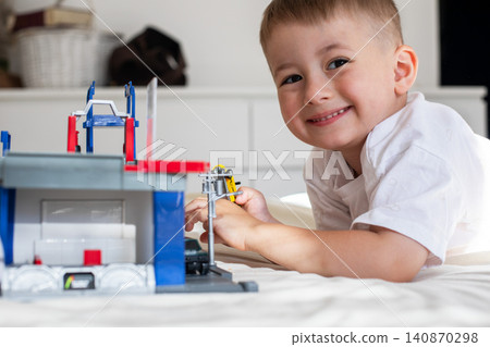 Cute young boy lying on a white blanket and focusing on playing with a toy garage, car repair set, and colorful scale model vehicles. Cute young boy lying on a white blanket and focusing on playing with a toy garage, car repair set, and colorful scale model vehicles. 140870298