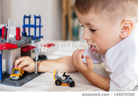 Cute young boy lying on a white blanket and focusing on playing with a toy garage, car repair set, and colorful scale model vehicles. Cute young boy lying on a white blanket and focusing on playing with a toy garage, car repair set, and colorful scale model vehicles. 140870300