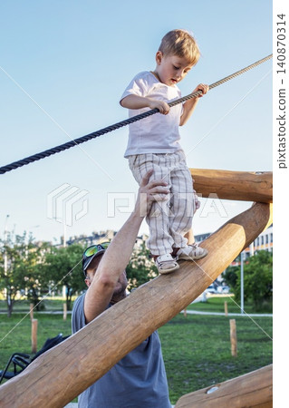 A caring father hands helps his little toddler boy walking safely on a wooden logs balance structure at outdoor city playground. A caring father hands helps his little toddler boy walking safely on a wooden logs balance structure at outdoor city playground. 140870314