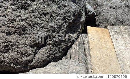 A top down horizontal perspective captures a massive porous volcanic rock boulder resting heavily against weatherbeaten wooden planks. The intense morning sunlight accentuates the rugged dark stone A top down horizontal perspective captures a massive porous volcanic rock boulder resting heavily against weatherbeaten wooden planks. The intense morning sunlight accentuates the rugged dark stone 140870596