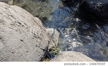 A high angle horizontal perspective captures clear clean water flowing gently between large textured river rock boulders. Bright morning sunlight reflects off the rippling water surface, creating A high angle horizontal perspective captures clear clean water flowing gently between large textured river rock boulders. Bright morning sunlight reflects off the rippling water surface, creating 140870597