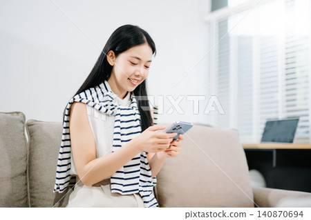 Happy Asian woman analyzing financial data documents and working on her laptop while sitting comfortably on a living room sofa. Happy Asian woman analyzing financial data documents and working on her laptop while sitting comfortably on a living room sofa. 140870694
