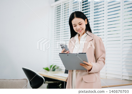 Happy Asian Businesswoman Reviewing Documents and Talking on Smartphone by Office Window in Modern Home Workspace Happy Asian Businesswoman Reviewing Documents and Talking on Smartphone by Office Window in Modern Home Workspace 140870703