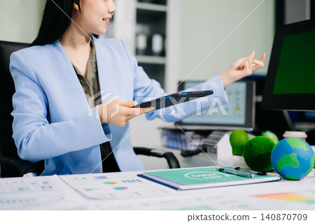 Smiling businesswoman holding a mini solar panel model at her office desk, surrounded by eco-friendly icons and net zero documents. Smiling businesswoman holding a mini solar panel model at her office desk, surrounded by eco-friendly icons and net zero documents. 140870709