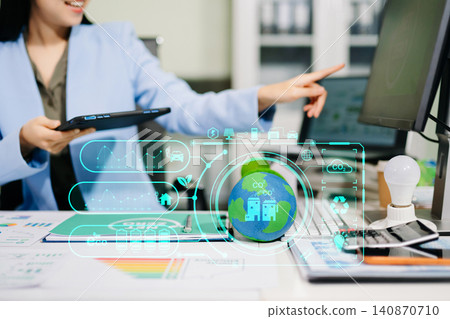 Smiling businesswoman holding a mini solar panel model at her office desk, surrounded by eco-friendly icons and net zero documents. 140870710