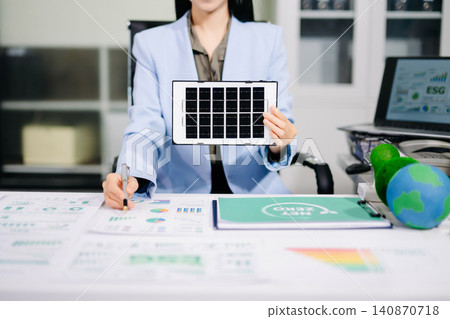 Smiling businesswoman holding a mini solar panel model at her office desk, surrounded by eco-friendly icons and net zero documents. 140870718