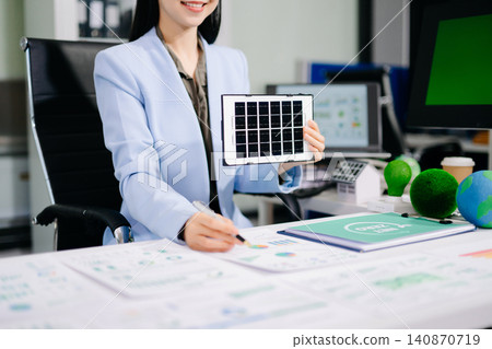 Smiling businesswoman holding a mini solar panel model at her office desk, surrounded by eco-friendly icons and net zero documents. 140870719