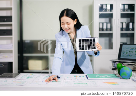 Smiling businesswoman holding a mini solar panel model at her office desk, surrounded by eco-friendly icons and net zero documents. Smiling businesswoman holding a mini solar panel model at her office desk, surrounded by eco-friendly icons and net zero documents. 140870720