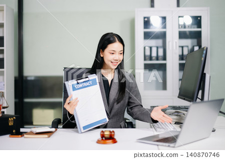Asian female lawyer holding a notebook and reviewing financial data charts on a computer monitor at a desk with a judge gavel. Asian female lawyer holding a notebook and reviewing financial data charts on a computer monitor at a desk with a judge gavel. 140870746