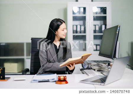 Asian female lawyer holding a notebook and reviewing financial data charts on a computer monitor at a desk with a judge gavel. Asian female lawyer holding a notebook and reviewing financial data charts on a computer monitor at a desk with a judge gavel. 140870748