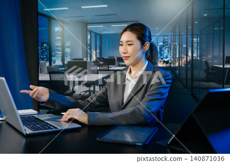 Asian businesswoman using a laptop and tablet in a modern night office. High-tech corporate workspace background. Ideal for business, technology Asian businesswoman using a laptop and tablet in a modern night office. High-tech corporate workspace background. Ideal for business, technology 140871036