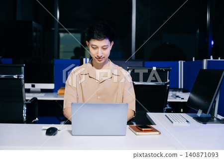 Happy Asian young man smiling while typing on his laptop at a modern corporate office desk at night. Happy Asian young man smiling while typing on his laptop at a modern corporate office desk at night. 140871093