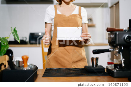 Close up of a female barista in a brown apron holding a white open sign behind a wooden cafe counter. 140871193