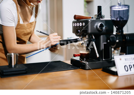 Female Asian barista using an espresso machine and taking notes on a clipboard in her coffee shop. Female Asian barista using an espresso machine and taking notes on a clipboard in her coffee shop. 140871205