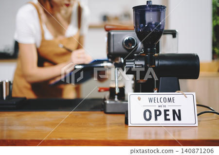 Barista writing order behind coffee machine and open sign on a warm wooden counter. Barista writing order behind coffee machine and open sign on a warm wooden counter. 140871206