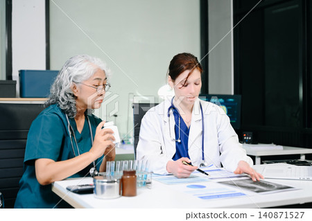 Senior and young female doctors reviewing data reports and a prescription bottle at a medical desk. 140871527