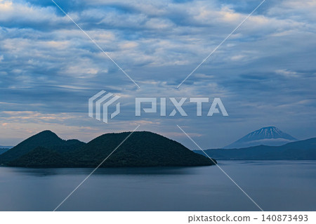 A dramatic view of Lake Toya, the lush Nakajima Island, and Mount Yotei stretching out beneath a sky filled with clouds. A dramatic view of Lake Toya, the lush Nakajima Island, and Mount Yotei stretching out beneath a sky filled with clouds. 140873493