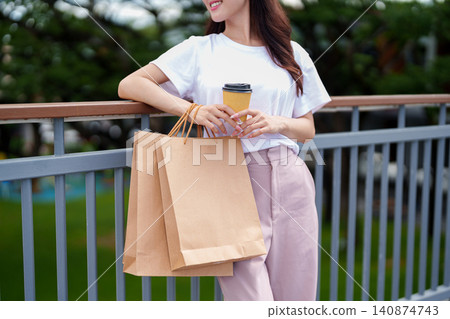 Young Asian woman holding coffee cup and shopping bags on outdoor terrace with natural bokeh background Young Asian woman holding coffee cup and shopping bags on outdoor terrace with natural bokeh background 140874743