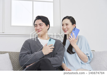 A portrait of a couple in their living room, holding a smartphone and looking directly at the camera. A portrait of a couple in their living room, holding a smartphone and looking directly at the camera. 140874784