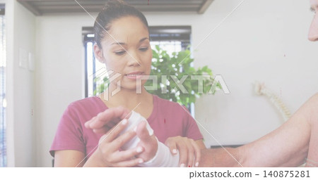 Wrapping caregiver in magenta top bandaging senior wrist with white bandage in clinic, spine model Wrapping caregiver in magenta top bandaging senior wrist with white bandage in clinic, spine model 140875281