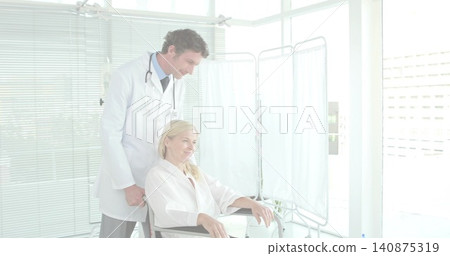 Leaning clinician in white coat examining patient in gown in wheelchair at clinic, stethoscope Leaning clinician in white coat examining patient in gown in wheelchair at clinic, stethoscope 140875319