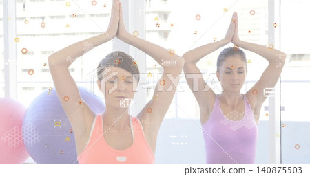 Two women doing arms-up pose in gym with city windows, balls, tech overlays, coral-lavender tanks 140875503