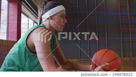 Sitting basketball player holding orange ball on wooden gym floor, in green jersey white headband Sitting basketball player holding orange ball on wooden gym floor, in green jersey white headband 140876022