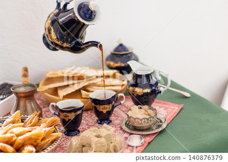 Pour coffee from a cobalt blue vintage decanter pot in cup. Sweets food on table honey baklava and halva on a patterned red tablecloth Pour coffee from a cobalt blue vintage decanter pot in cup. Sweets food on table honey baklava and halva on a patterned red tablecloth 140876379