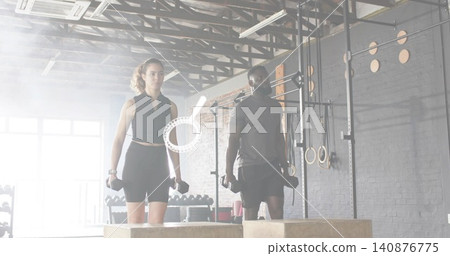 Standing holding hex dumbbells, woman and man in black gear at crossfit gym behind plyo boxes Standing holding hex dumbbells, woman and man in black gear at crossfit gym behind plyo boxes 140876775