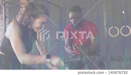 Pedaling woman in black tank gripping air bike handles at gym, coach gesturing in red shirt Pedaling woman in black tank gripping air bike handles at gym, coach gesturing in red shirt 140876782