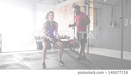 Swinging female client in shorts, tank top doing low squat in gym, trainer coaching, battle ropes Swinging female client in shorts, tank top doing low squat in gym, trainer coaching, battle ropes 140877158