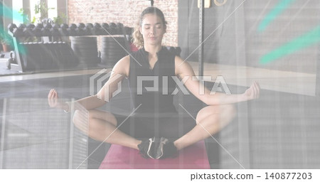 Sitting woman meditating on pink mat in gym, wearing black top, leggings, dumbbell rack nearby Sitting woman meditating on pink mat in gym, wearing black top, leggings, dumbbell rack nearby 140877203