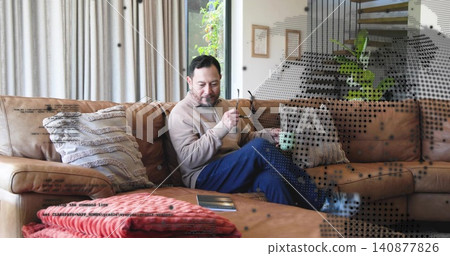 Sitting Asian male in tan pullover, blue trousers on leather sofa, holding glasses with green mug Sitting Asian male in tan pullover, blue trousers on leather sofa, holding glasses with green mug 140877826